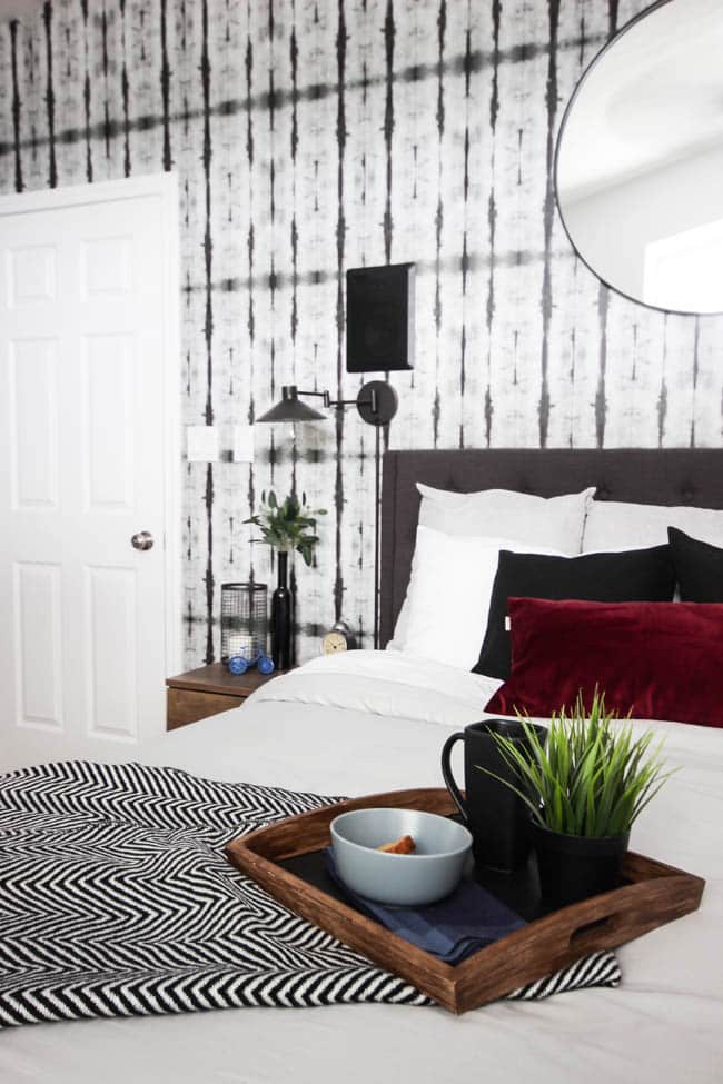 A beautiful Modern Master Bedroom Renovation Reveal! Gorgeous bold wallpaper, black, white and grey tones alongside wooden accent furniture. Ikea cabinets with a sliding barn door. And a white brick fireplace with a dark wooden mantel. Touches of blue and deep red finish the space perfectly. Beautiful transformation! 