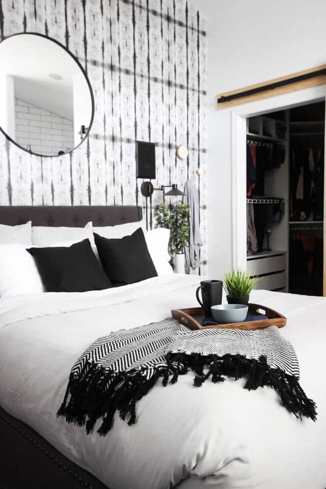 A beautiful Modern Master Bedroom Renovation Reveal! Gorgeous bold wallpaper, black, white and grey tones alongside wooden accent furniture. Ikea cabinets with a sliding barn door. And a white brick fireplace with a dark wooden mantel. Touches of blue and deep red finish the space perfectly. Beautiful transformation! 