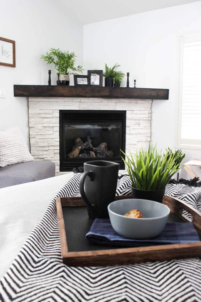 A beautiful Modern Master Bedroom Renovation Reveal! Gorgeous bold wallpaper, black, white and grey tones alongside wooden accent furniture. Ikea cabinets with a sliding barn door. And a white brick fireplace with a dark wooden mantel. Touches of blue and deep red finish the space perfectly. Beautiful transformation!