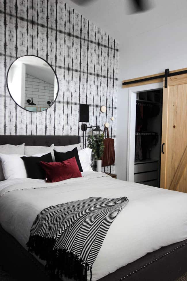 A beautiful Modern Master Bedroom Renovation Reveal! Gorgeous bold wallpaper, black, white and grey tones alongside wooden accent furniture. Ikea cabinets with a sliding barn door. And a white brick fireplace with a dark wooden mantel. Touches of blue and deep red finish the space perfectly. Beautiful transformation! 