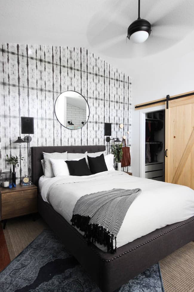 A beautiful Modern Master Bedroom Renovation Reveal! Gorgeous bold wallpaper, black, white and grey tones alongside wooden accent furniture. Ikea cabinets with a sliding barn door. And a white brick fireplace with a dark wooden mantel. Touches of blue and deep red finish the space perfectly. Beautiful transformation! 