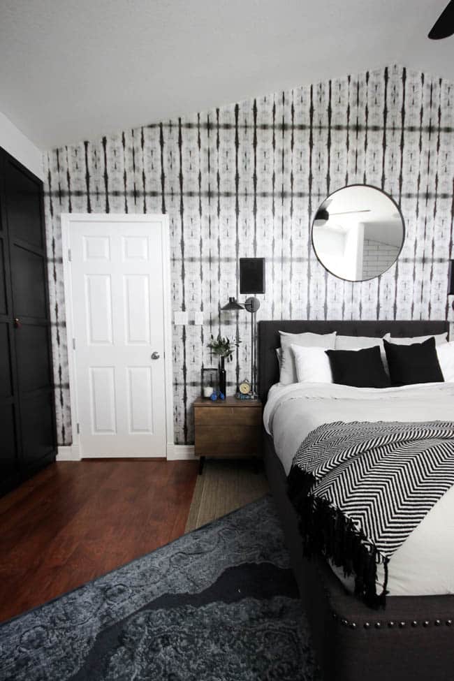 A beautiful Modern Master Bedroom Renovation Reveal! Gorgeous bold wallpaper, black, white and grey tones alongside wooden accent furniture. Ikea cabinets with a sliding barn door. And a white brick fireplace with a dark wooden mantel. Touches of blue and deep red finish the space perfectly. Beautiful transformation! 