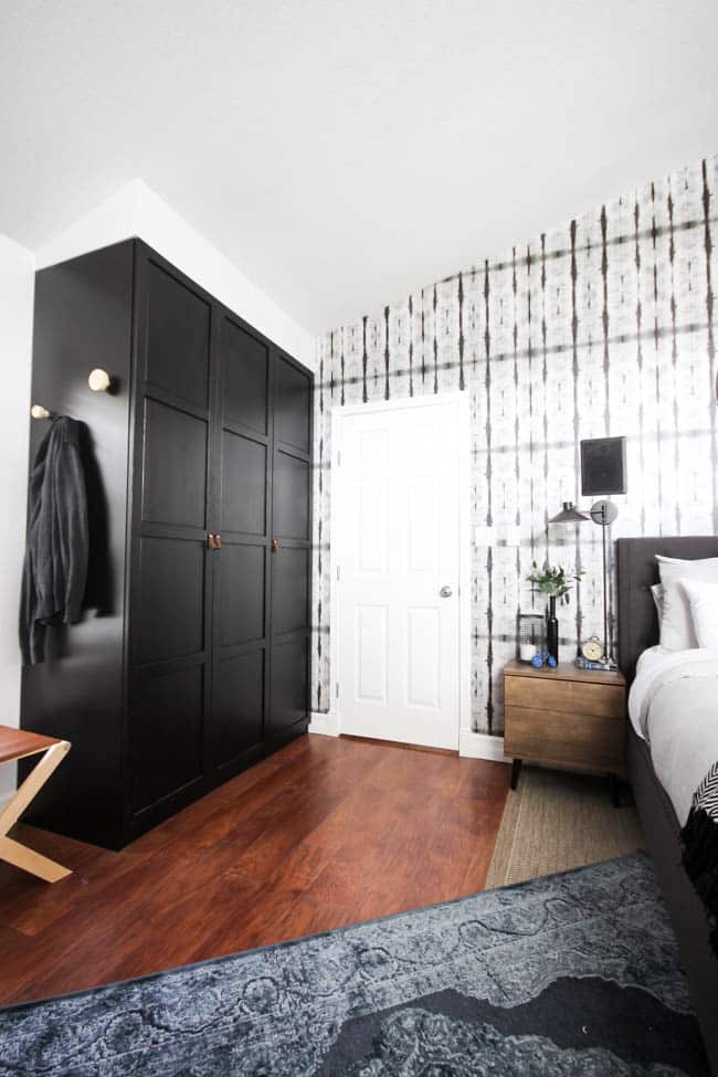 A beautiful Modern Master Bedroom Renovation Reveal! Gorgeous bold wallpaper, black, white and grey tones alongside wooden accent furniture. Ikea cabinets with a sliding barn door. And a white brick fireplace with a dark wooden mantel. Touches of blue and deep red finish the space perfectly. Beautiful transformation! 