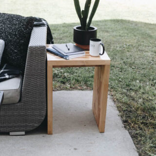 Outdoor wood side table next to outdoor sofa with plant, coffee mug, and notebooks and pen