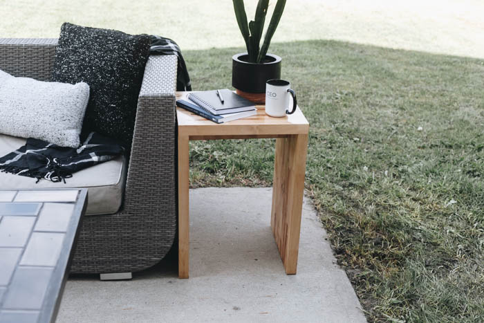 Outdoor wood side table next to outdoor sofa with plant, coffee mug, and notebooks and pen
