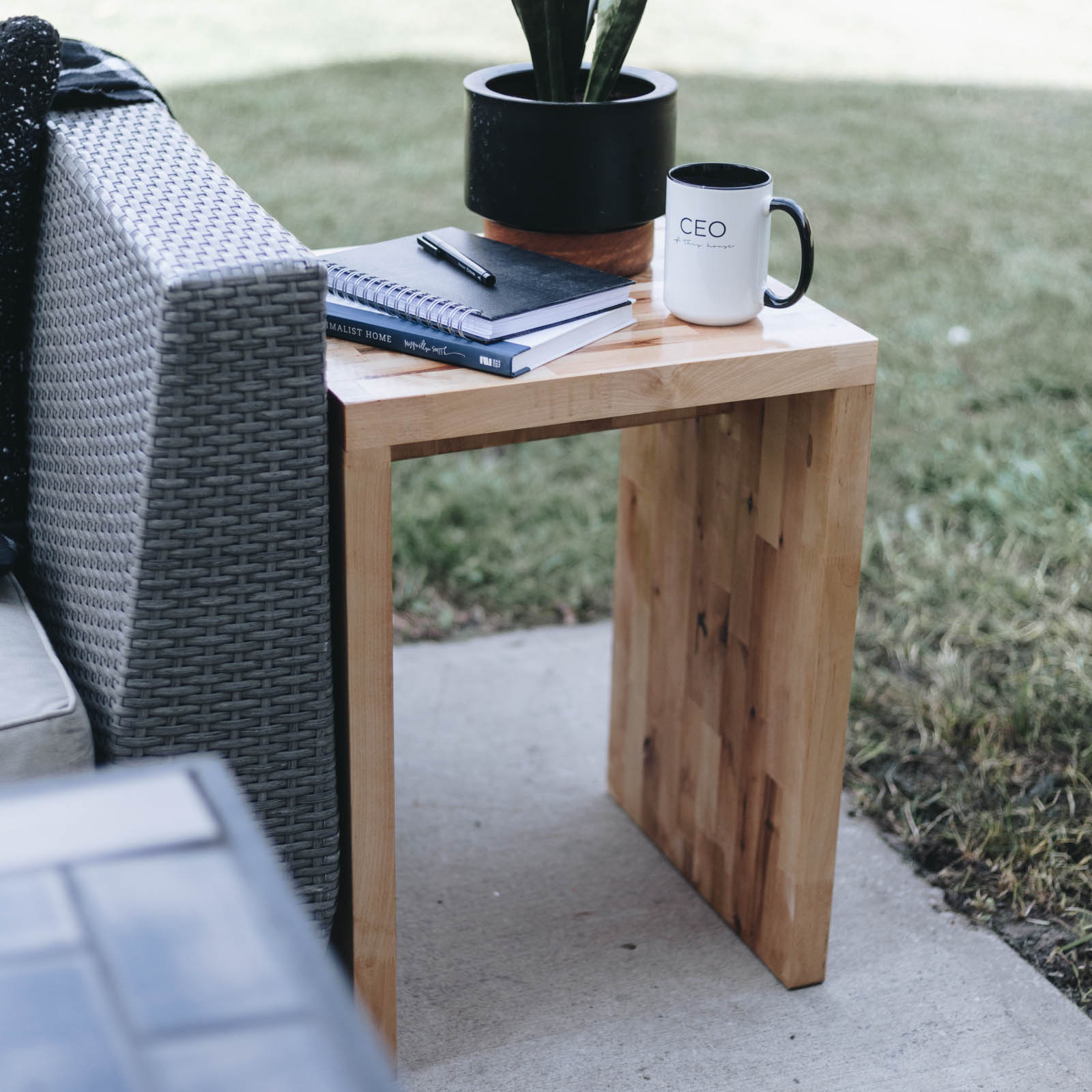 Outdoor wood side table next to outdoor sofa with plant, coffee mug, and notebooks and pen